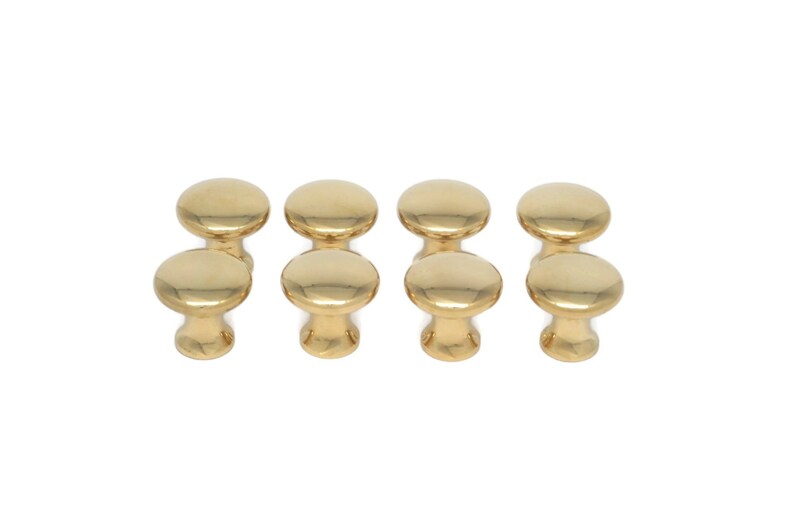 May include: Eight gold-colored cabinet knobs with a rounded top and a short, cylindrical base. The knobs are arranged in two rows of four, against a white background. These knobs are suitable for furniture.