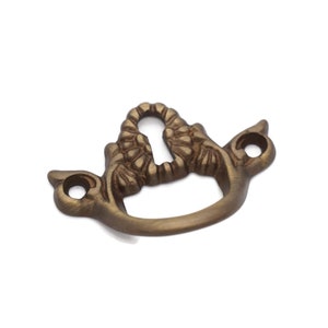 May include: A gold-toned metal keyhole cover with a decorative floral design. The cover has a curved shape and two small holes on the sides.
