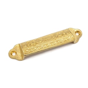 May include: A gold-toned metal drawer pull with an ornate design. The pull has a rectangular shape with rounded edges and a decorative pattern.