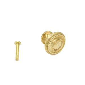 May include: A gold-toned cabinet knob with a round, slightly raised design. It comes with a matching gold screw.