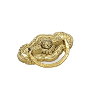 May include: A gold-toned ornate cabinet pull with a decorative ring handle. The pull has a floral design and a central round knob.