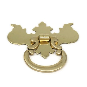 May include: A gold-toned metal drawer pull with a decorative, ornate design. The pull features a ring-shaped handle with a decorative backplate.