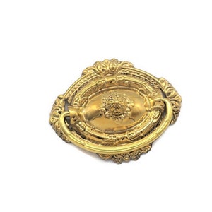 May include: A gold-toned ornate oval-shaped cabinet pull with a decorative ring handle.