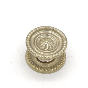 May include: A gold-toned cabinet knob with a decorative, raised, floral design. The knob has a beaded edge.