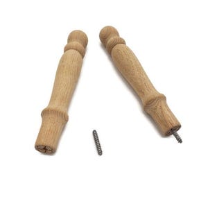 May include: Two unfinished wooden furniture legs with a decorative turned design. A small metal screw is positioned between the legs. The legs are light brown and appear to be new.