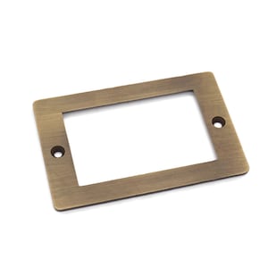 May include: A brass-colored rectangular frame with a smaller rectangular opening in the center. The frame has a brushed finish and two small holes on the sides.