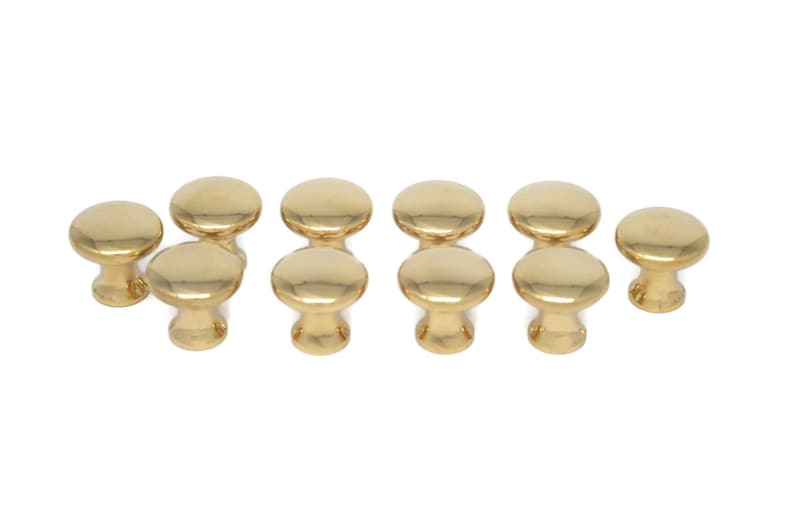 May include: A collection of ten shiny, gold-colored cabinet knobs. Each knob has a rounded top and a short, cylindrical base. The knobs are arranged in two rows, showcasing their polished finish and simple design. Ideal for furniture upgrades.