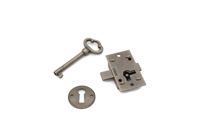 May include: Antique-style hardware set featuring a key, a keyhole cover, and a lock mechanism. The metal pieces have a tarnished, bronze finish, suggesting an aged aesthetic. The key has a decorative handle, and the lock has a visible keyhole.