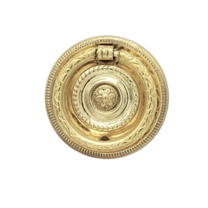 May include: A gold-toned, circular cabinet pull with a decorative design. The pull features concentric circles with detailed patterns and a hinged ring. The hardware is likely for furniture or cabinetry.