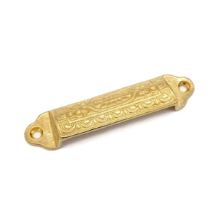 May include: Gold-toned metal drawer pull with an ornate design. The pull has two holes for mounting.
