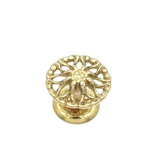 May include: A gold-toned metal cabinet knob with a decorative, circular design. The knob has a raised, intricate pattern that resembles a flower or star.