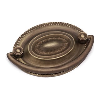 May include: An antique brass cabinet pull with an oval-shaped plate and a decorative handle. The plate has a raised, textured design and a beaded edge.