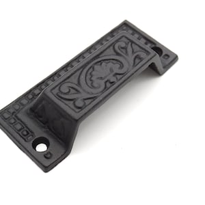 May include: Black cast iron door pull with an ornate floral design. The pull has a rectangular shape with rounded edges and two screw holes.