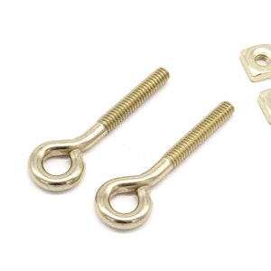 May include: Two gold-toned eyelet screws with square nuts. The screws have a threaded shaft and a loop at the end. The nuts are square and have a hole in the centre.
