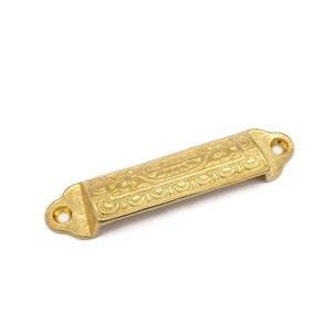 May include: Gold-toned metal drawer pull with an ornate design. The pull is rectangular with rounded edges and has two holes for mounting.