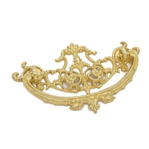 May include: Ornate gold-colored drawer pull with a curved, symmetrical design. The pull features intricate floral and scrollwork details, with two rounded end pieces for attachment. A decorative hardware element.