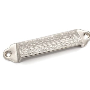 May include: A silver-toned, ornate drawer pull with a rectangular shape and a decorative design. The pull has two holes for mounting.