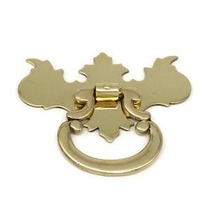 May include: A gold-toned metal drawer pull with a decorative, ornate design. The pull features a ring-shaped handle with a decorative backplate.