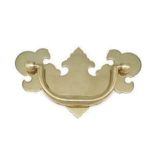May include: A gold-toned metal cabinet pull with a decorative, ornate design. The pull has a curved, D-shaped handle and two decorative finials on either side.