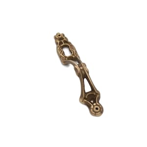 May include: Ornate, antique-style bronze drawer pull with a decorative openwork design. The pull features a central oval opening and two mounting holes at the base. Ideal for furniture restoration or adding vintage charm.