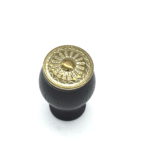 May include: A black wooden knob with a gold metal decorative top. The top has a circular design with a raised center.