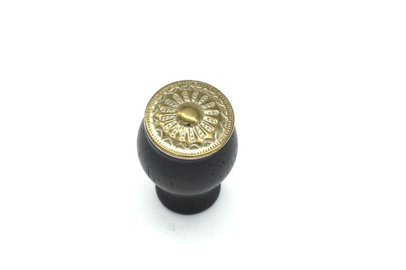 May include: A black wooden knob with a gold metal decorative top. The top has a circular design with a raised center.