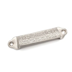 May include: A silver-toned, rectangular drawer pull with an ornate, embossed design. The pull has two holes for mounting.
