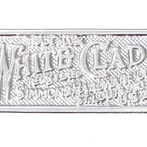 May include: A silver-colored metal plate with the words "White Clad" in embossed lettering. The plate has a textured background and the text "Simmons Hardware Co. St. Louis, USA". Two holes are on either end.