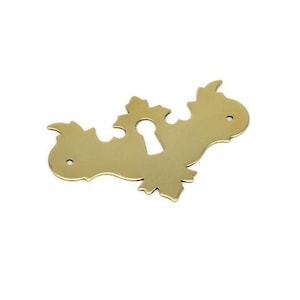 May include: A brass keyhole cover with a decorative, ornate design. The cover is shaped like a stylized leaf with a keyhole in the center.
