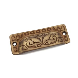 May include: A brass drawer pull with an ornate floral design. The pull is rectangular with rounded edges and has a small hole on each side for mounting.