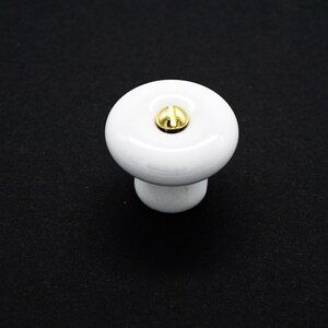 May include: White ceramic cabinet knob with a gold metal screw. The knob is round with a slightly raised top.