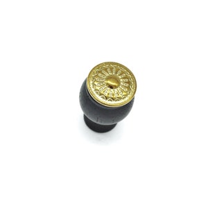 May include: A black wooden cabinet knob with a gold metal decorative top. The top has a circular design with a raised center.