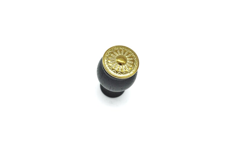 May include: A black wooden cabinet knob with a gold metal decorative top. The top has a circular design with a raised center.