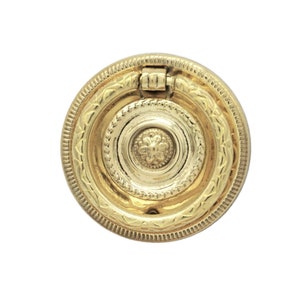 May include: A gold-toned metal ring pull with a decorative, ornate design. The ring pull has a circular shape with a raised center and a detailed border.