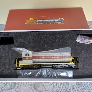 May include: A model train locomotive in a white box with a black foam insert. The locomotive is grey with a maroon stripe and yellow accents. It says "ERIE LACKAWANNA" on the side and "421" on the front.