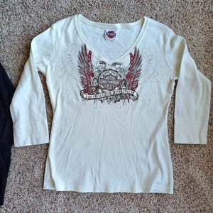 May include: Cream-coloured long-sleeved shirt with a V-neck. The shirt features a Harley-Davidson Motorcycles graphic in red, grey, and black on the front. The graphic includes wings and the Harley-Davidson logo.