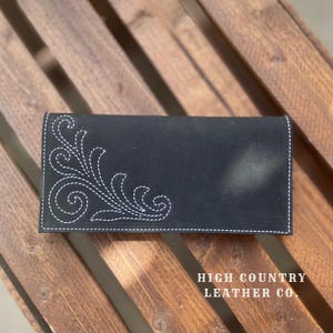 Peut inclure: Portefeuille en cuir noir avec des coutures décoratives blanches. Le portefeuille présente un motif floral détaillé sur le devant et le texte "HIGH COUNTRY LEATHER CO." en bas à droite.