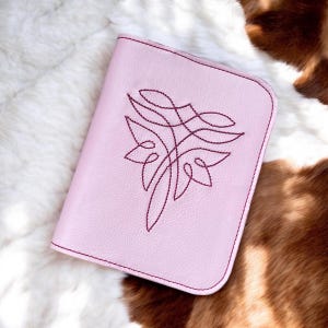 Op de afbeelding: Een roze leren notitieboek met een bordeauxrood geborduurd ontwerp van een gestileerde bloem.
