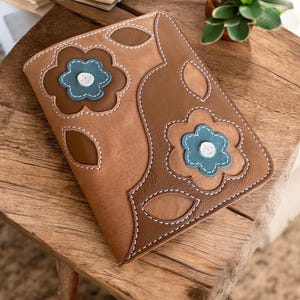 Op de afbeelding: Een bruine leren notitieboekomslag met een bloemenpatroon. De bloemen zijn uit het leer gesneden en hebben een tealblauwe kern. De omslag heeft een gestikte rand.