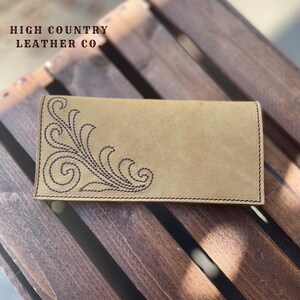 Peut inclure: Un portefeuille en cuir beige avec un motif floral brodé marron foncé dans le coin. Le portefeuille est rectangulaire et porte le texte "HIGH COUNTRY LEATHER CO" en haut. Le portefeuille est posé sur une surface en bois.