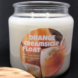 May include: A glass jar candle with a white label that says "Orange Creamsicle Float" and "Taylor Home Goods Handmade Soy Candle". The label also has a picture of an orange creamsicle float.