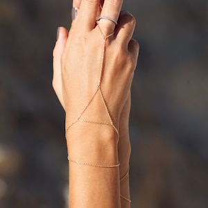 Puede incluir: Un brazalete de cadena dorada con un diseño delicado. El brazalete se lleva en la muñeca y se extiende por el brazo, terminando en un anillo en el dedo.