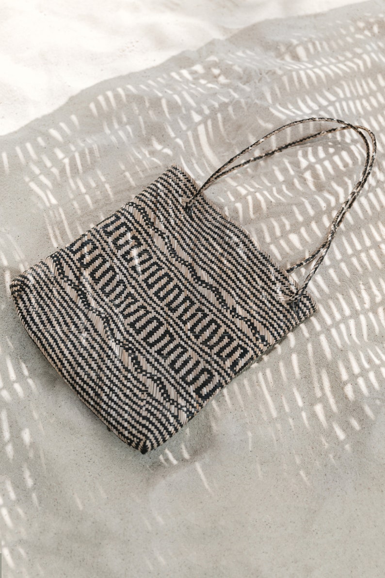 May include: A black and white woven tote bag with a geometric pattern. The bag is lying on a white sandy surface with shadows.