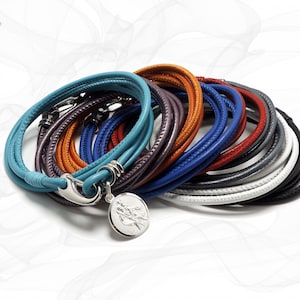 Könnte beinhalten: Ein Set aus bunten Lederarmbändern mit einem silbernen Anhänger. Die Armbänder sind in verschiedenen Farben übereinander gelegt, darunter Blau, Lila, Orange, Rot und Weiß.