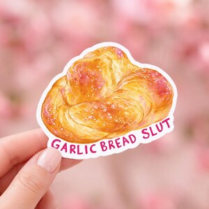 May include: A die-cut sticker featuring a golden-brown, braided garlic bread illustration with a glossy finish. The text GARLIC BREAD SLUT is printed in bold, dark pink capital letters along the bottom edge of the bread-shaped sticker design.