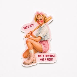 May include: A die-cut sticker featuring a blonde person with a pink hair bow, wearing a white collared shirt and pink shorts with tan boots, holding a wooden baseball bat. The text reads KNEECAPS ARE A PRIVILEGE, NOT A RIGHT in red letters.