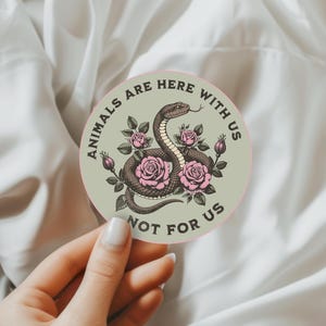 May include: Round sticker with a snake illustration surrounded by pink roses. The sticker has the text "ANIMALS ARE HERE WITH US NOT FOR US" in a circular pattern around the edge. The background is a light green color.