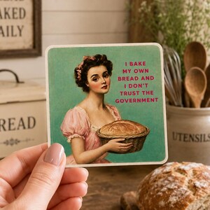 May include: A square coaster featuring a vintage-style illustration of a person in a pink dress holding a basket of bread. The text reads: I BAKE MY OWN BREAD AND I DON'T TRUST THE GOVERNMENT. The background is a solid teal color with a matte finish.