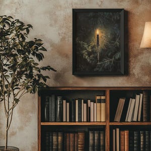 May include: A framed print of a candle burning in a dark forest setting. The candle is on a candlestick and surrounded by pine branches. The image has a dark, moody feel.