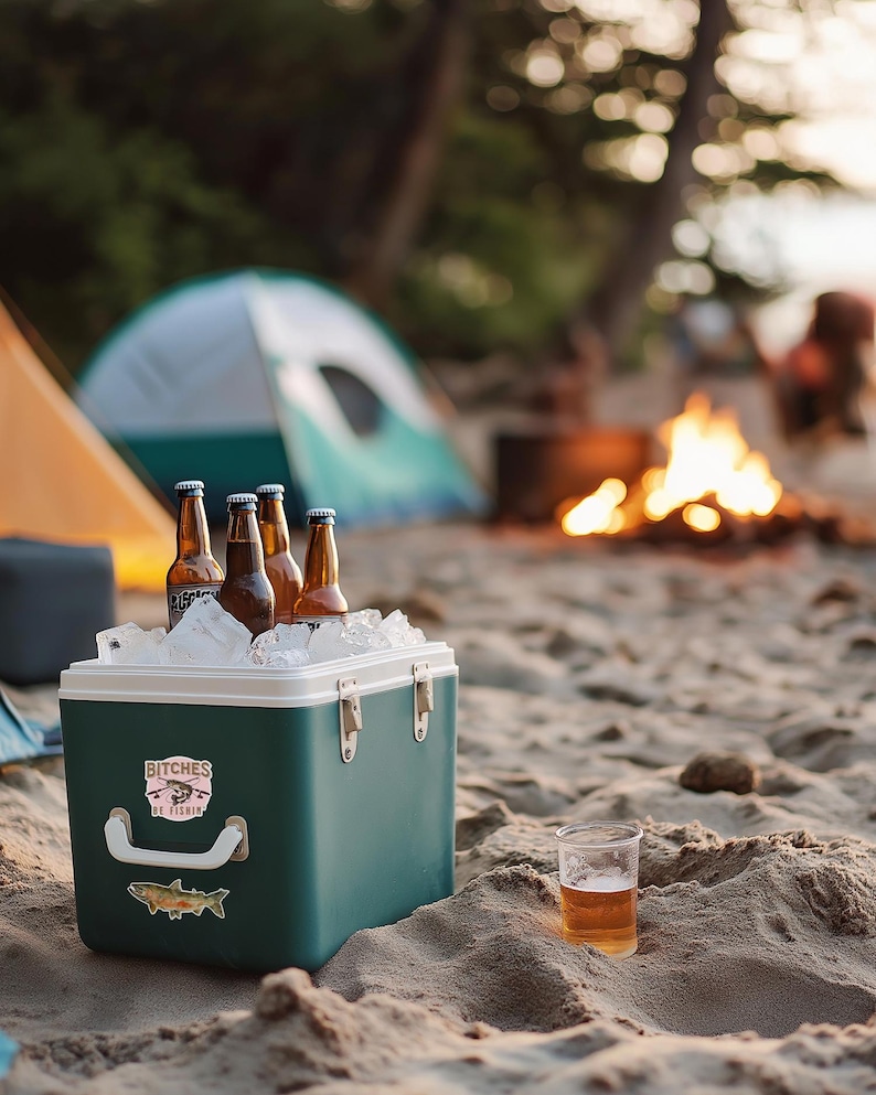May include: A green cooler with a sticker that says "Bitches Be Fishin'" and a fish illustration. The cooler is filled with ice and beer bottles. A glass of beer sits on the sand in the foreground.